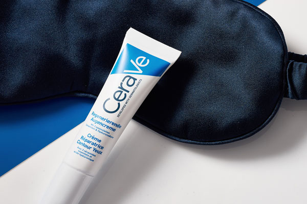 Die CeraVe Regenerierende Augencreme mit pflegendem Jojobaöl lehnt an einer schwarzen Schlafmaske